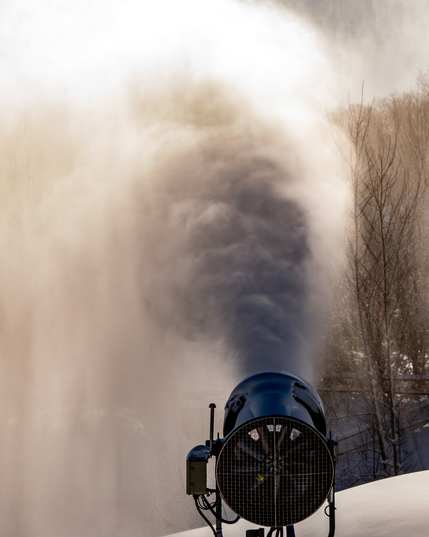 A cylindrical machine with large fan is pointing away from the viewer and torrent of fine snow is being blown of the presumably snow making machine, into the air creating a mist lighted by the bright sun
