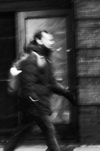 Black and white photo of a man walking on the sidewalk in the snow. He is passing by a door that frames him 