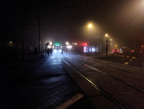 A tram in the dark. A slight haze from the lights of the tram with black silhouettes of people passing in front of it. 