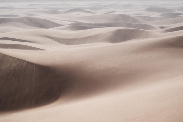 Soft, wind-sculpted sand dunes in layered beige tones, with rippled textures, sweeping shadows, and a faint line of footprints crossing the midground.