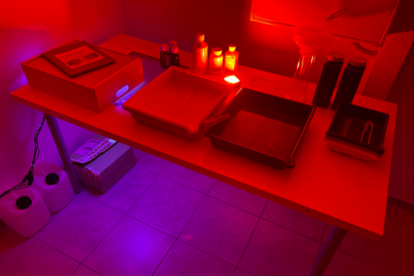 Long exposure photo of a basic DIY darkroom setup in my basement, featuring an old table with a bunch of photo trays (different sizes), bottles of chemicals for developing prints, a wooden box converted into an UV exposure unit for contact printing