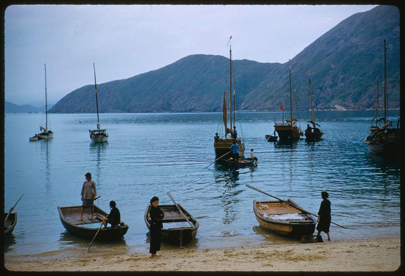 A group of small fishing boats are tied up on a beach, with the water and mountains in the background. There is one larger sailboat at sea out towards the horizon. Some people stand or sit around some of these boats while others walk by carrying paddles or poles; two men appear to be pulling their boat onto land using oars. The sky has light cloud cover over it, but it seems mostly bright and sunny.
