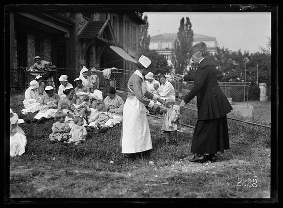 The image depicts a black and white scene with individuals dressed in what appears to be early 20th-century attire. A woman wearing a dark coat, skirt, hat, and shoes is extending her hand towards another person who is handing something small to a child. Surrounding them are adults and children seated on the grass; many of these figures wear hats or caps typically associated with healthcare settings from that era.

The backdrop includes buildings resembling institutional structures, possibly hospitals or institutions for care, along with some greenery and fencing in the background. The overall composition suggests an outdoor visit by a dignitary named Miss Anne Maxwell to this institution, where she is greeting individuals who may be patients or residents of such facilities at the time.