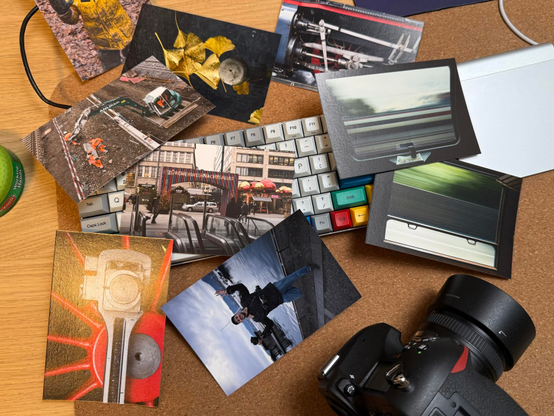 A flat lay of photographs on a wooden surface, featuring images of construction scenes, urban settings, and nature. A DSLR camera and a colorful keyboard are also visible among the scattered photos.