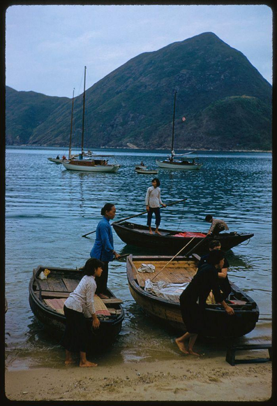 Several people are at the shore, handling their small boats while larger sailboats float nearby on a calm bay in Hong Kong. A person wearing black and squatting next to one of these motorless rowboats is assisting another person who stands upright as they prepare for departure from land. The other three individuals also appear to be preparing or disembarking their vessels. In the background, hills rise up against an overcast sky.
