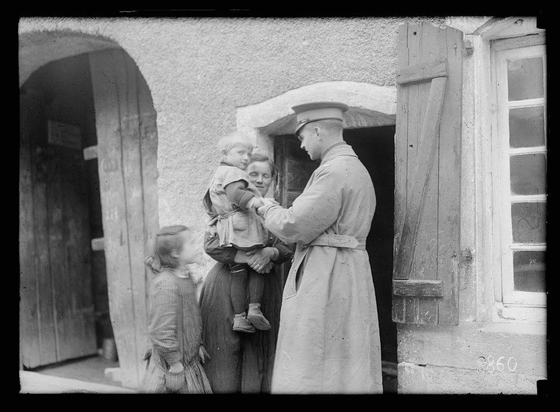 The image is a black and white photograph depicting four individuals standing in front of an old building with wooden shutters. On the left, there are two children dressed in period clothing; one child appears to be about three years old and wearing a cap, while the other seems younger, around 18 months, also wearing a hat but not holding anything. Between them is a woman who looks towards her right side where an adult male figure stands with his back partially turned toward us as he addresses or examines something held in his hands.

The man wears a light-colored coat and cap typical of military-style uniform from the early 20th century, suggesting that he might be part of some official capacity. His attire is complemented by glasses and seems to hold an object for inspection with both hands. The surrounding environment hints at rural France during wartime given the historical context provided in the caption.

The setting suggests a scene where medical care or assistance was being administered outdoors due possibly to lack of indoor facilities, which aligns with activities carried out by organizations like ARC (American Red Cross). There's no direct indication that it’s Christmas time; however, such events could have been part of larger humanitarian missions during wartime periods.