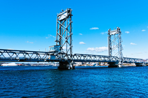 A bridge has a low-slung deck over the water with two tall towers in the center under a blue sky with just a few white clouds in the distance.
