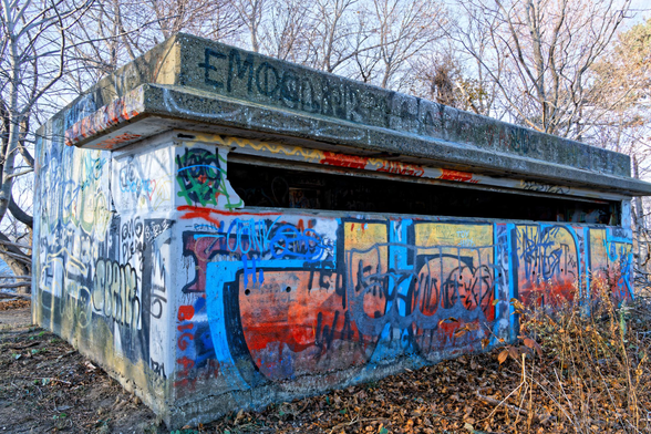 A concrete  bunker with colorful red, yellow and blue graffiti has a narrow slot to look out of and has winter-phase trees that aren't very tall behind it.