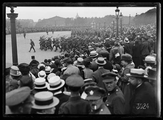 The image depicts a historical event where American troops, brought in from the Chateau Thierry front to participate in an event on July 4th, are seen marching across what appears to be Place de la Concorde. The photo captures them walking in formation with rifles at rest and bayonets fixed. They move past a large crowd of spectators who have gathered around, some wearing hats typical of early 20th-century fashion.

The setting is open-air on a cloudy day with the iconic structures visible in the background, including what seems to be the National Assembly building (Palais Bourbon) at one end and possibly other historical landmarks. The audience consists mainly of adults dressed in formal attire, indicating that this was likely an important public event.

A distinctive feature in the foreground is a tall lamppost with ornate details, characteristic of Parisian streetscape architecture from around 1900. There's also text "3024" visible on the right side of the image which could be indicative of its cataloging or archival number within a collection.

The overall ambiance suggests an occasion where national pride and military display are central themes during this period in history, possibly symbolizing unity and patriotism as seen through participation in such events.