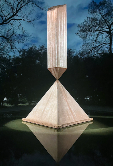 A towering, illuminated sculpture made of weathered, rust-colored material featuring a pyramid base with an inverted obelisk balanced precariously point-to-point on its apex. The obelisk's top is jagged and broken. The sculpture stands in a reflecting pool at night, casting a mirror image on the water, surrounded by dark tree silhouettes against a cloudy, deep blue sky.