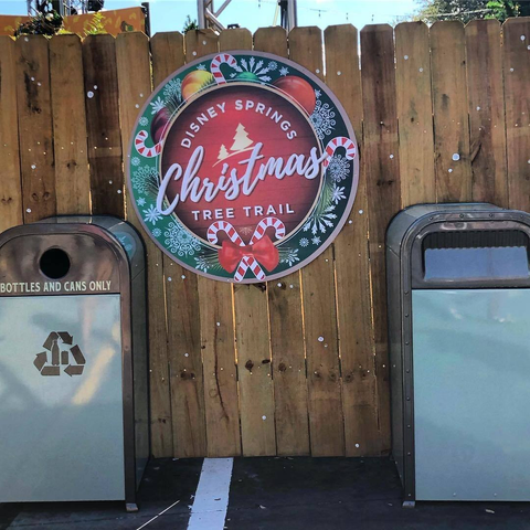 Two metal trash cans stand side by side in front of a wooden fence at Disney Springs. The left can is labeled “Bottles and Cans Only” with a recycling symbol, while the right can is plain. Above them, a round festive sign reads “Disney Springs Christmas Tree Trail” with candy cane accents and greenery. Small white foam specks, resembling snow, are scattered on the fence, part of the park’s artificial “snoap” effect.