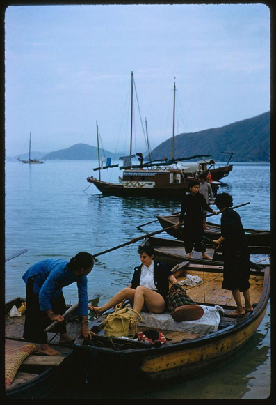 an image depicting a boat with people on it. the photograph was taken by toni frissel and is titled "frissel's hong kong assignment". this particular picture appears to have been captured during an assignment for sports illustrated in 1959, featuring views of various boats along with mountains in the background.