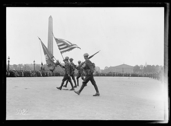 The image depicts a historical scene with several individuals in military uniforms marching across an open field. They are carrying flags and guns, suggesting they may be part of a parade or demonstration related to the American armed forces. The background features notable landmarks such as the Washington Monument, which signifies that this event is taking place in Washington, D.C. A large crowd can also be seen gathered around the perimeter of the open field, watching the procession.

The black and white photograph indicates it was taken during a time when color photography was not prevalent or used for documenting events like this. The image carries historical significance as it captures a moment related to American military history, potentially from World War I era given the attire and context provided by the caption.