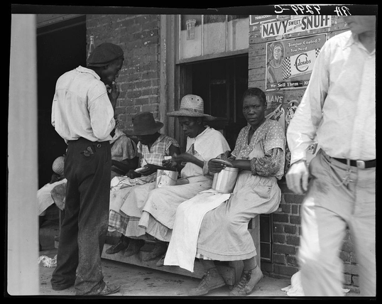 The image is a black and white photograph depicting four individuals seated outside on a bench. Three of them appear to be women wearing light-colored, long-sleeved dresses with aprons over their garments; the fourth individual seems male in casual work attire, including pants and shoes that suggest manual labor or fieldwork. The man stands beside them, facing away from the camera while holding what appears to be a can or cup near his mouth, possibly drinking. They are situated outside of an establishment adorned with various signs advertising products like "Navy Sweet Snuff" and "Lane Labs," suggesting it might be a café or diner.

One woman is dressed in a darker dress, standing beside the others on the bench while holding what seems to be a cup or tin can. The women seated are engaged with their hands; one holds an object that appears to be money, perhaps counting bills as she sits next to another who also has some coins visible near her hand.

The men and women wear casual clothing appropriate for outdoor activity but do not seem to have heavy jackets or protective gear commonly associated with agricultural work. The environment suggests a historical period where manual labor was prevalent, possibly in agriculture given the style of dress suitable for fieldwork among them.