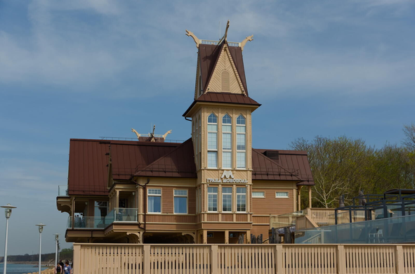 The image features a large, ornate building with a brown roof and white trim. The structure is topped with two spires that reach towards the sky. The building has multiple windows on its facade, adding to its grandeur. A small balcony can be seen on one side of the building. On the facade there are words “Grand Monopole” in cyrillic letters In front of the building, there are several people walking around.

The scene is set against a backdrop of trees and a body of water, creating a serene atmosphere. The sky above is clear blue with scattered clouds, suggesting a bright and sunny day.