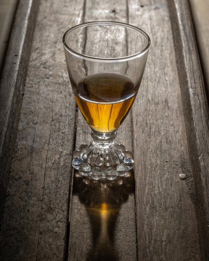 A photograph of a glass containing amber-colored whiskey on an aged, unfinished wooden surface, lit from behind and casting a shadow with a caustic highlight the color of the whiskey in front of the glass. 