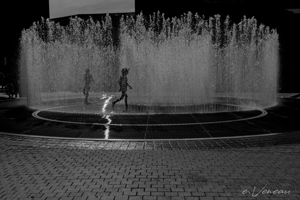 A ray of sunlight falls on rising water jets in the city: white of the jets on a black background. Two children are playing in the middle: black silhouettes against a white background of water jets.