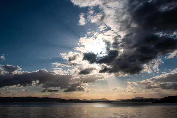 Ein Abendhimmel zur blauen Stunde an der Applecrossbay mit Blick auf Raasay und Isle of Skye: Dunkle Wolken schieben sich vor die Sonne, der Horizont ist warm golden-bräunlich gefärbt, das Wasser schimmert. 

An evening sky at twilight at Applecross Bay with a view of Raasay and the Isle of Skye: dark clouds drift across the sun, the horizon is a warm golden brown, and the water glistens. 