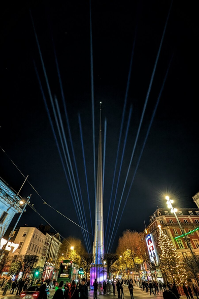 A low-angle night photograph capturing the Spire of Dublin towering over O'Connell Street. Multiple beams of intense blue light project upwards from the base of the needle-like monument, converging into the dark sky. The street below is lively with pedestrians and a green double-decker bus, while the surrounding buildings are illuminated by warm streetlamps and festive Christmas tree lights on the right.