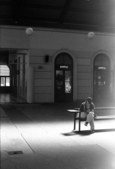 A person sits alone on a bench in a sunlit, empty indoor space. They are reading or looking at something in their hands. The background features arched doorways and storefronts, creating a tranquil atmosphere. The image is presented in black and