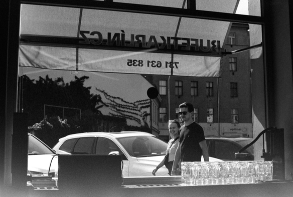 The image shows two people walking outside a building, viewed through a window. A row of glass mugs is visible in the foreground, while cars are parked outside. The scene is captured in black and white, giving it a classic feel.