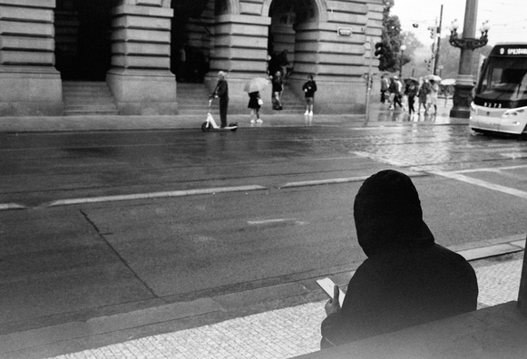 A person in a black hoodie sits on a bench, looking at a phone. The scene is rainy and captures a street with pedestrians and a tram in the background. The image is in black and white, emphasizing the urban environment.