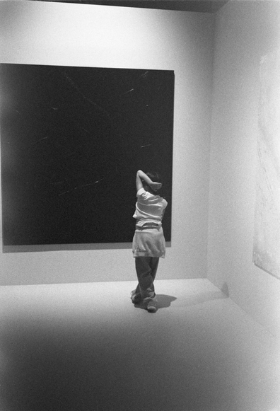 A figure is seen standing in an art gallery, facing a large black canvas. The person has their arms raised behind their head, seemingly contemplating the artwork. The image is in black and white, emphasizing the minimalist setting.