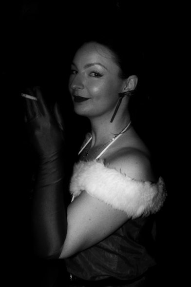 Black and White photo of a woman sitting with her side to the camera. She is dressed in a fancy looking old style dress and holding a cigarette. Smiling at the camera.