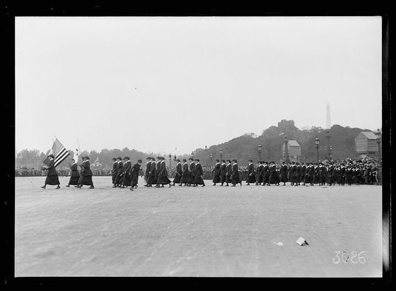 The image is a black and white historical photograph depicting an organized procession. The setting appears to be a large open public space, possibly during the early 20th century given its monochrome nature.

In the foreground, we see several women dressed in long dark dresses with sashes around their waists, walking on what looks like grass or dirt ground. They are all wearing hats and some have handbags slung over one shoulder. A few of these individuals carry flags; notably, a woman is holding an American flag.

Behind them follows a group of people who seem to be dressed in military uniforms with caps and dark coats, suggesting they may belong to the Army Medical Corps or another branch related to public health services during wartime periods when such corps were prominent. Some of these figures are carrying instruments that resemble bagpipes.

In the background stands an audience composed primarily of women in similar attire as those leading; their heads suggest hats or headscarves which was common for middle-class women's wear at that time. The setting seems festive and celebratory, possibly a parade given the organized nature of everyone’s movements and decorations like flags being displayed prominently.

The photograph is labeled with "3026" in the bottom right corner indicating it may be part of an archive or collection series by Lewis Hine who was known for his [...]