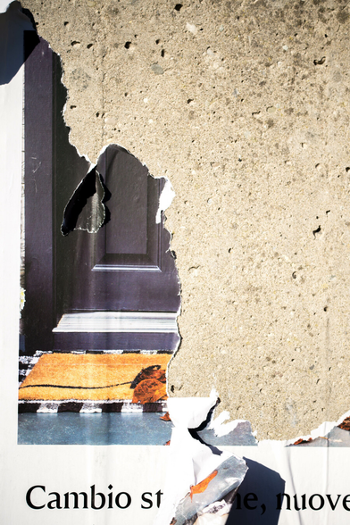 Photo of a brightly sunlit, mostly torn poster. The top right is all pockmarked concrete, the bottom left is the remains of a poster that has a dark door above a black and white-edged mat, underneath which are the words 'CAMBIO' then something obscured by a flap of torn poster then ' NUOVE'. Where the poster is torn, the bright sunlight creates dark shadows.