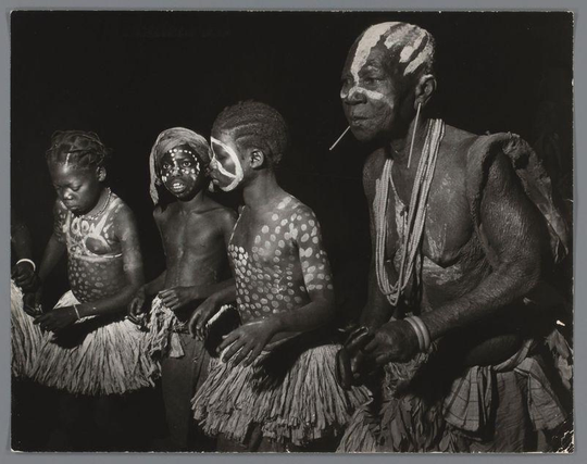 The image depicts a group of individuals, likely participating in a cultural or ceremonial event. They are adorned with traditional body paint and patterns that include white circles on their skin, suggesting some form of tribal identity or celebration. The person to the right stands out due to his larger-than-life appearance with similar painted face markings. He is wearing multiple strands of beads around his neck and has visible arm rings. All individuals wear grass skirts, which are part of traditional African attire.

The photograph captures a moment that exudes cultural richness, possibly from an indigenous community's gathering or performance. The black-and-white nature of the image adds to its historical feel, indicating it might be taken in the late 20th century given the style and clothing seen here. No discernible modern elements are present; instead, there is a clear focus on tradition and heritage.

The background is dark, which serves to highlight the subjects even more prominently against the contrast provided by their painted skin and attire. This emphasis suggests that the photo aims to showcase these individuals in all of their cultural splendor without any external distractions. The composition directs attention toward the unity among the group members while also inviting viewers into a moment frozen in time, offering a glimpse into another culture's pr [...]