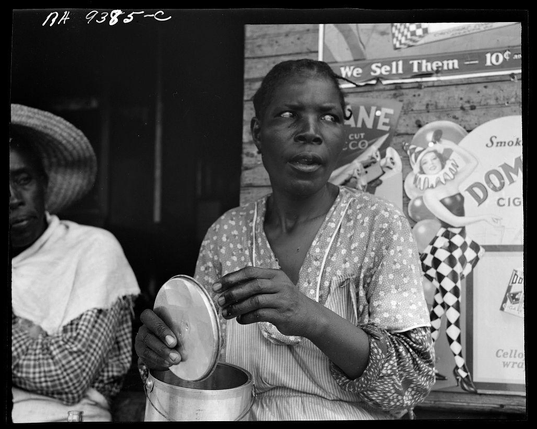 The photograph captures a moment with two individuals who appear to be engaged in an activity or conversation, possibly outdoors. The focus is on the person seated and holding what seems to be a large metal container or utensil, which they are examining closely.

This individual has dark skin tones, wears patterned clothing that includes light colors and stripes, and appears somewhat contemplative as she looks towards something off-camera with her eyes wide open. Her attire suggests practicality for outdoor work, possibly related to manual labor given the context provided by additional signage in the background.

Beside this person sits another individual partially visible on the left side of the frame. This second person is wearing a white and black patterned garment that covers their head entirely with what looks like a hat or protective covering from sun exposure. Their posture indicates they are also partaking in the activity, but not as prominently involved.

The background contains various elements including signage such as advertisements for products like cigarettes labeled "DOMINO" and other indistinct texts hinting at commercial activities. The setting seems to be an urban environment with a rustic or makeshift feel given the textures and materials visible on the walls behind them. There's also visual clutter, indicating a busy or bustling location.

Overall, thi [...]