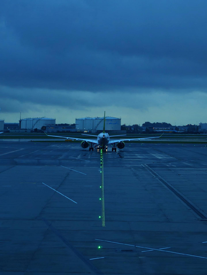 A photo showing an airplane taxiing on a wet airport tarmac at dusk. A line of green lights leads straight toward the aircraft. Large fuel or storage tanks are visible in the background.
