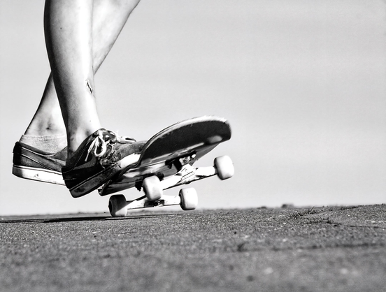Das Bild zeigt in einer Schwarz-Weiß-Aufnahme die Beine und Füße einer Person, die auf einem Skateboard steht und sich offenbar fortbewegt. Der Fokus liegt auf der dynamischen Bewegung und den Details der abgenutzten Schuhe sowie des Skateboards. Die Szene spielt sich auf einem rauen, asphaltierten oder betonierten Untergrund ab, der im Vordergrund scharf abgebildet ist, während der Hintergrund unscharf und hell gehalten ist, was den Blick auf das Hauptmotiv lenkt. Die gekreuzten Beine und die Position des Boards vermitteln den Eindruck von Balance und Action.