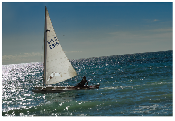 Querformat: ein kleiner Sportkatamaran mit zwei Rümpfen segelt von rechts nach links in spätem Winterlicht.  Man sieht eine helles weisses Segel  und eine dunkel angezogen Person, glitzerndes blaues Wasser, schiefer Meereshorizont.
