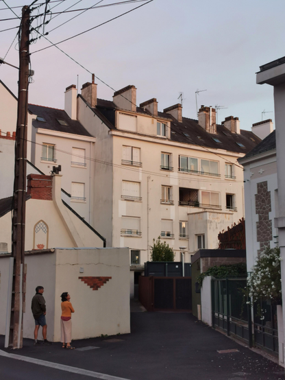 Façade d'un immeuble au soleil couchant, intégré dans un tissu urbain dense. A l'entrée de la ruelle devant l'immeuble, un couple observe quelque chose.