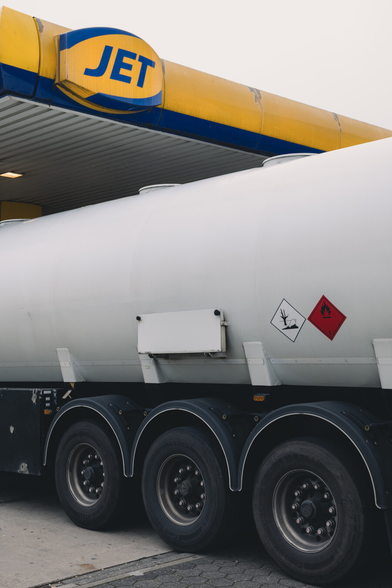Un camion-citerne blanc est garé sous l’auvent d’une station-service JET, identifiable par son enseigne jaune et bleue. On ne voit qu'une partie de l'auvent, et qu'une partie centrale du camion. Sur le côté de la citerne, deux pictogrammes de danger sont visibles. Le bas de l’image montre les roues arrière du camion. Les passages de roues forment des arcs de cercles qui tranchent avec les lignes droites de l'auvent et du camion