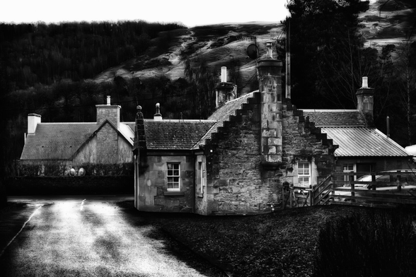 Traditionelle Steinhäuser mit markanten Schornsteinen und Stufengiebeln stehen im Vordergrund einer schottischen Landschaft. Die Schwarz-Weiss-Darstellung betont die Texturen der Steinmauern und die winterliche Ästhetik der Umgebung, mit einer feuchten Strasse und dunklen Hügeln im Hintergrund.