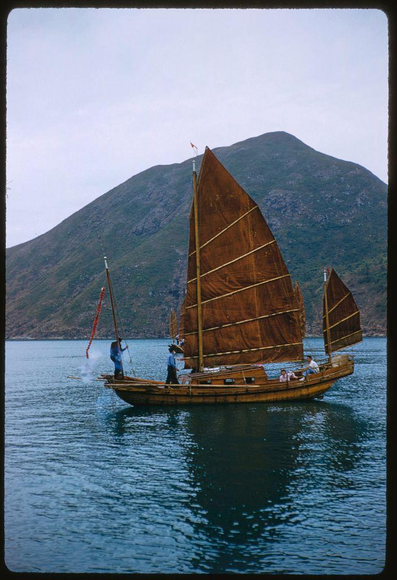 A wooden sailboat with large brown sails is on a calm body of water near the shore, where two people are standing and working. The mountainous landscape in the background features lush greenery covering its slopes and appears to be under an overcast sky. This scene was captured by renowned American photographer Toni Frissell during her assignment for Sports Illustrated in 1959