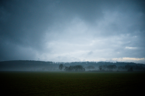 Wiese Bäume in der ferne. Nebel. Hügel. Ein Mast ganz klein am Berg/Hügel. Wolken. Strucktur in den Wolken.