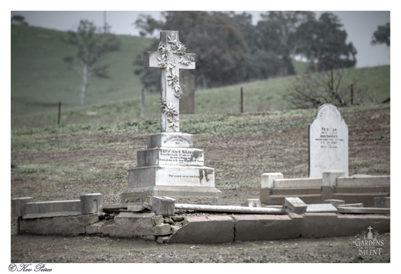 A detailed, weathered, carved stone cross stands prominently in the foreground of a cemetery in a rural setting.  The cross is decorated with carved vines and flowers, and rests on a stacked, tiered pedestal with inscriptions.   Surrounding the base are broken, low stone or concrete borders. In the mid-ground, a simple, rounded white headstone is visible to the right, and the background consists of sloping, brown and green grassy hills under an overcast sky.