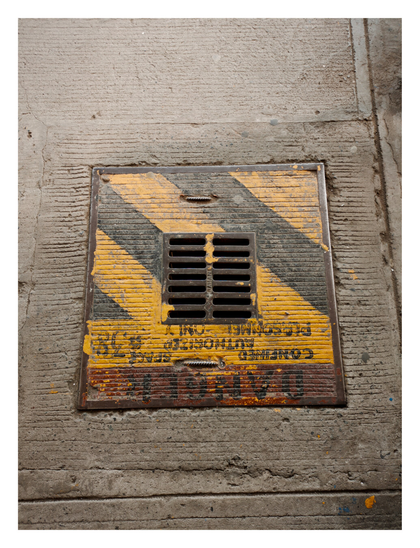 A heavily weathered metal drain or access cover embedded in rough concrete pavement. The cover is painted with diagonal yellow and black hazard stripes and features a central ventilation grate. Faded, inverted safety warning text, including "CONFINED SPACE" and "AUTHORIZED PERSONNEL ONLY," is visible on the surface. - Google Gemini Latest