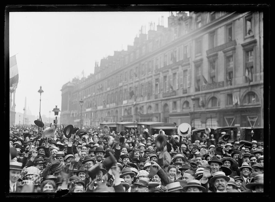 The image depicts a large gathering of people on a street, seemingly in celebration or protest. The crowd is dense with individuals wearing various styles of hats and headwear such as bowlers, fedoras, and wide-brimmed hats typical of early 20th-century fashion. Many are raising their hats or waving them enthusiastically, which suggests excitement or triumph.

Behind the people, there's a backdrop of large buildings that resemble European architecture from around the time period depicted in black-and-white photography—likely late 19th to mid-20th century given the style and state of wear on the structures. The street is lined with lampposts, and what appears to be trolley cars or trams are visible further down the road.

The overall atmosphere conveyed by this image is one of high emotion, possibly related to a significant public event occurring at Rue Royale near Place de la Concorde, as referenced in the alt text provided.