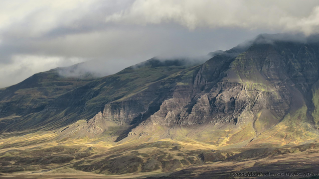 A colour photo of a mountainside of brown rock with sharp ridges running down the flanks. Low sunlight has caught the front edge of these and shadowed behind them, producing a clear outline of the geology. The tops are in cloud and the sky is grey. The foreground is rounded hills created by landslips from the peaks above.