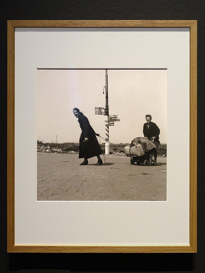 Een zwartwit foto van twee vrouwen tijdens de hongerwinter in Nederland. De rechtervrouw duwt een kinderwagen vol met spullen die kunnen worden geruild voor eten, de linkervrouw trekt met een touw de kinderwagen. In het midden staat een paal met ouderwetse plaatsaanduidingen. Op de achtergrond zie je bomen en huizen. Deze foto is stiekem door de fotograaf Cas Oorthuys gemaakt.