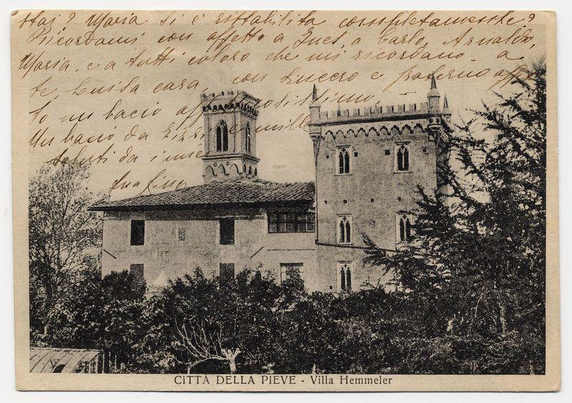The image displays a postcard featuring an old, monochromatic photograph of the Villa Hemmeler in Città della Pieve. The architectural style is reminiscent of medieval or renaissance design with its pointed arch windows and towers that suggest defensive characteristics typical for structures from those periods.

There's handwritten text on the upper part of the image which seems to be personal notes, possibly by someone associated with the villa, but it appears faded in some parts making full transcription challenging. Below this script is printed text stating "CITTA DELLA PIEVE - Villa Hemmeler", identifying both the location and subject.

The photograph captures a side view of what looks like an imposing building surrounded by trees which obscure much of its lower facade, creating shadows across it. The details within the structure's design are somewhat obscured due to the age or quality of the print but one can discern tall narrow windows characteristic of such buildings' defensive architecture and some intricate detailing on the walls.

Overall, this image exudes a historical ambiance with an air of antiquity that could interest those in history, architecture or who have connections to Città della Pieve.