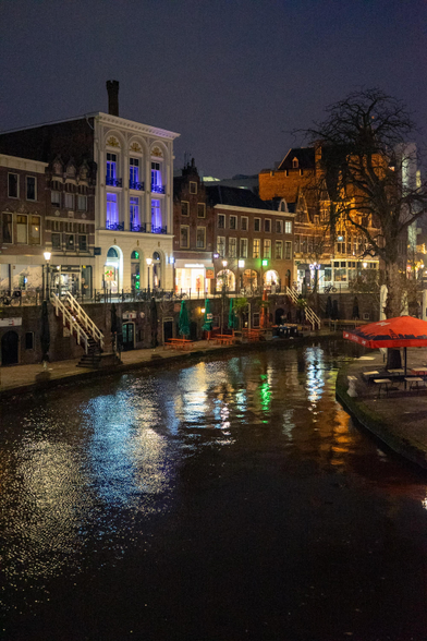 This image captures a nighttime scene of a canal in an urban area.

The canal, which is the central focus, is flanked by buildings on both sides. The water reflects the lights from the buildings and street lamps, creating a shimmering effect on the surface. On the left side, there are several multi-story buildings with illuminated windows, some of which have decorative architectural details. The building closest to the canal has a staircase leading down to the water.

On the right side, there are more buildings, some with outdoor seating areas covered by large umbrellas. Trees, some bare and others with sparse foliage, line the canal, adding to the urban landscape. The overall atmosphere is calm and serene, with the lights from the buildings and street lamps casting a warm glow on the scene. The sky is dark, indicating it is nighttime.

