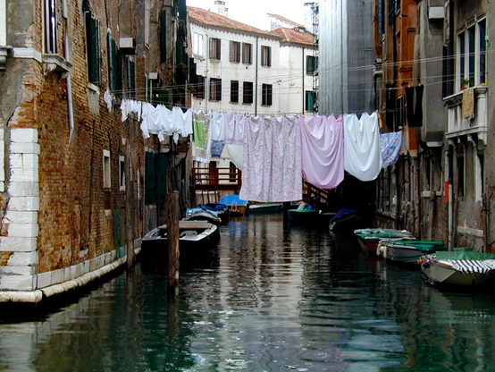 A colour picture looking along a small Venetian canal lined by domestic houses. There are small boats in the canal and washing lines draped across,  maybe 10 metres up, with bedsheets etc hanging out to dry - over the filthy canal water!
