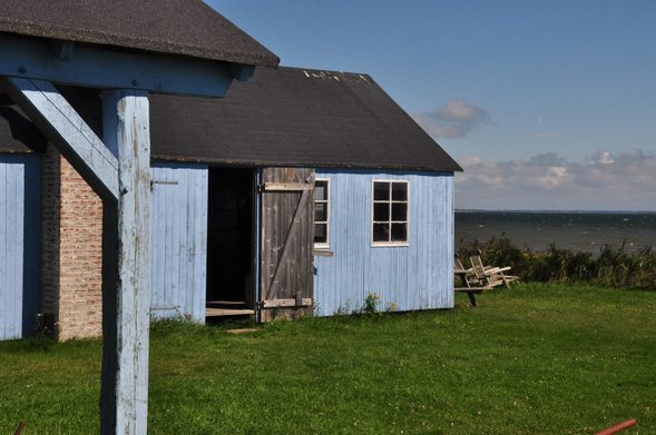 Der kleine himmelblau gestrichene Museumshof in Jütland ist einladend, aber nicht überlaufen. Mittig eine weit geöffnete Holztür, die eins der beiden weissen Sprossenfenster zur Hälfte verdeckt. Der Himmel setzt die Farbe des Hofs am Horizont fort und verläuft zum oberen Bildrand ins azurblau. Das Meer erscheint in der Ferne graublau und ist im Uferbereich aufgewühlt graubraun.  