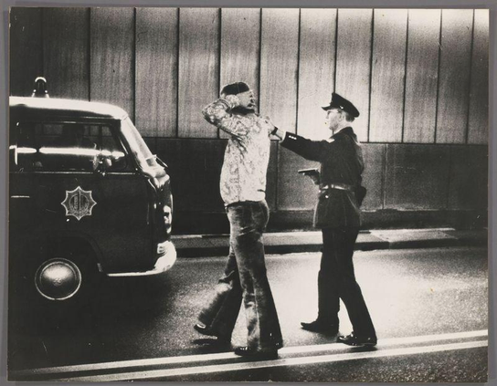 The image depicts a high-contrast black and white scene with two figures in the foreground. On the left, an individual stands beside a police vehicle, their hand raised near their face as if adjusting glasses or holding something to their mouth. The person is dressed casually, wearing what appears to be denim pants and possibly a patterned shirt.

On the right side of the image, another figure is standing in profile view towards us, facing away from the camera. This individual seems to be clad in police attire, complete with a hat, jacket, trousers, and boots—indicating an authoritative or law enforcement role.

The backdrop suggests an urban setting during dusk or dawn, given its muted lighting. The vehicle's emblem is visible on the side of the car door; however, it lacks clear detail that could be used to identify specific brands or models from a distance. Notably absent are any discernible features such as license plates, windows, or other identifying markers.

The composition and styling hint at an older photograph, possibly mid-20th century based on fashion styles and vehicle design elements. The image captures a moment of apparent confrontation or arrestation between the civilian and law enforcement figure in front of what might be their police car.